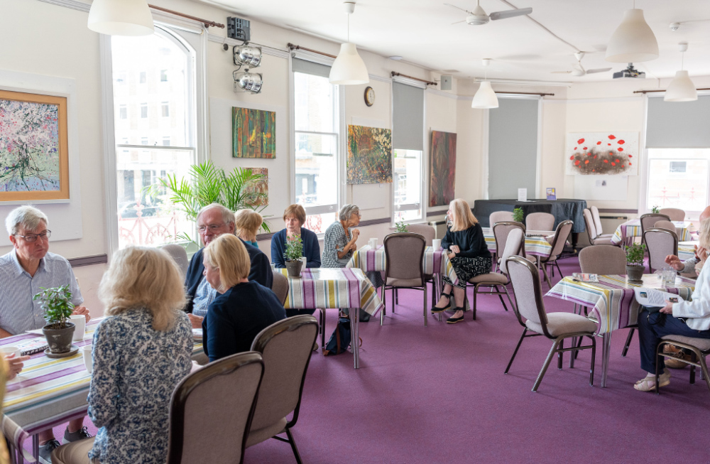 Groups of people are sat talking at different tables. The room has a purple carpet, white walls with artwork on display and large sash windows.