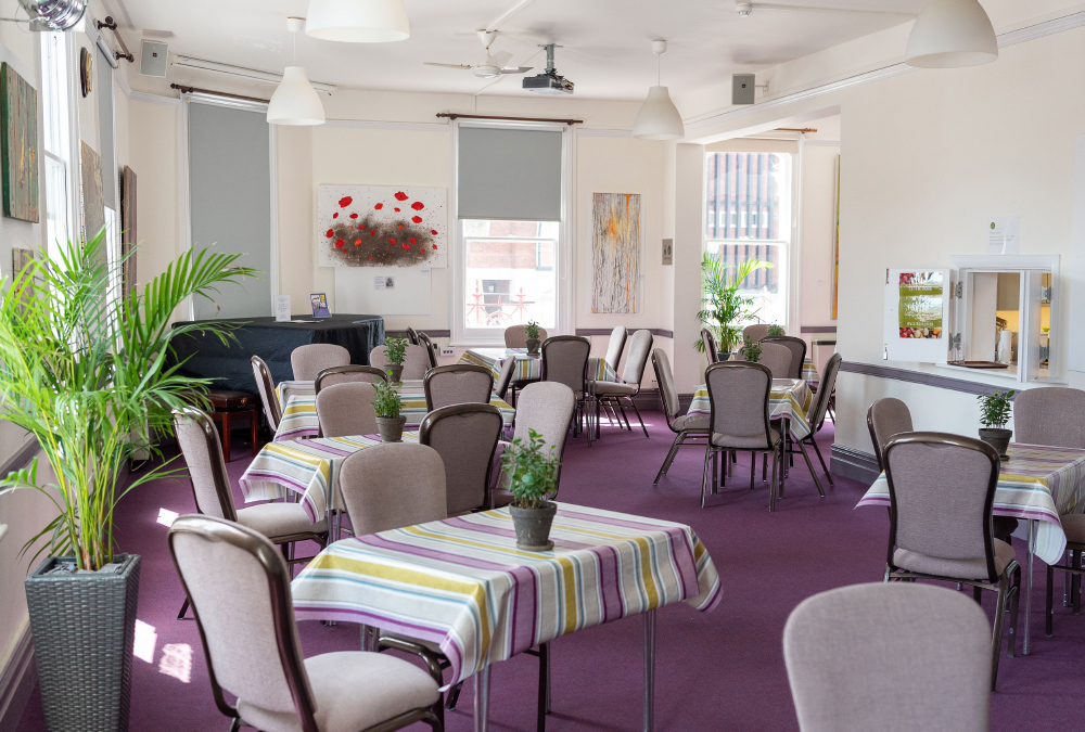 View of the room which is filled with grey chairs and tables covered with a purple, yellow and white striped tablecloth. There is a large, green palm in the foreground and artwork on the walls.