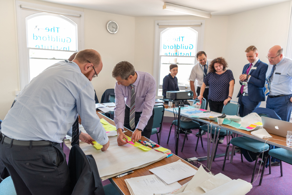 The Conference Room: view of 8 people having a meeting. 3 people are stood in the foreground at a table looking at information laid out on the table. Four others are stood in the background doing the same.