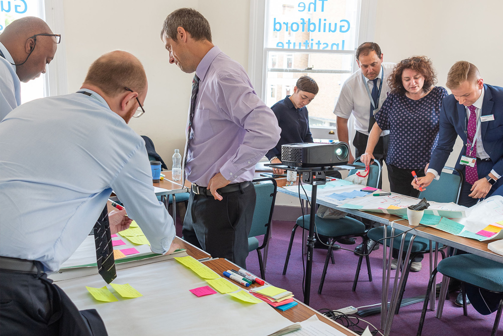 The Conference Room: view of 8 people having a meeting. 3 people are stood in the foreground at a table looking at information laid out on the table. Four others are stood in the background doing the same.