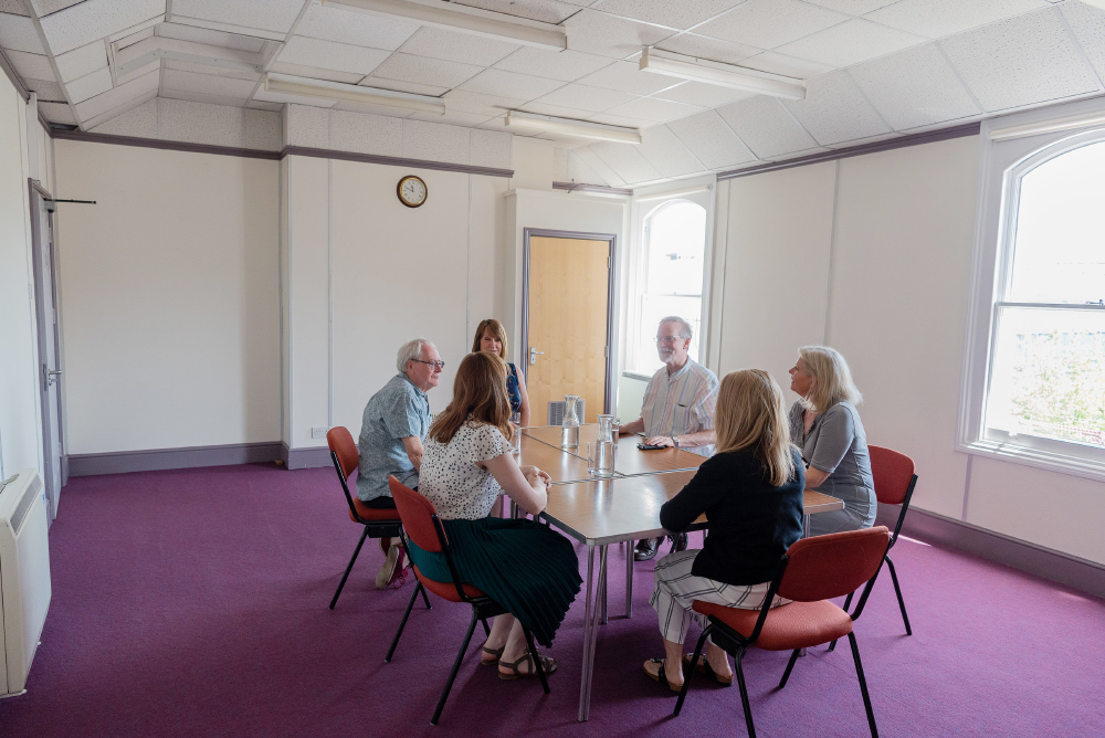 The Lecture Room: six people having a meeting, sat on red chairs around a wooden table. The walls are white with a purple trimming and the carpet is purple.