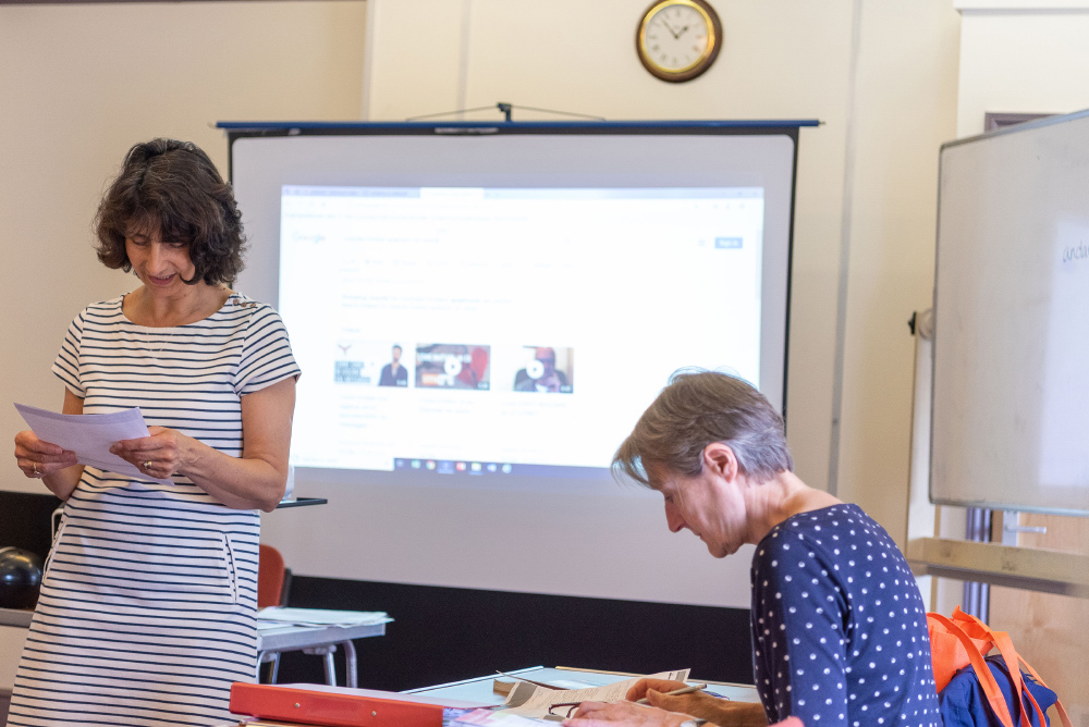 The Lecture Room: One woman stood up wearing a black and white striped dress, reading notes. A second woman wearing a navy and white polka dot top is sat reading. There is a large screen in the background projecting an image. 