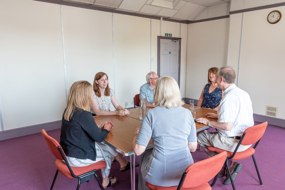 The Lecture Room: 6 people having a meeting. They are sat on red chairs around a wooden table. The walls are white with a purple trim and the carpet is also purple.