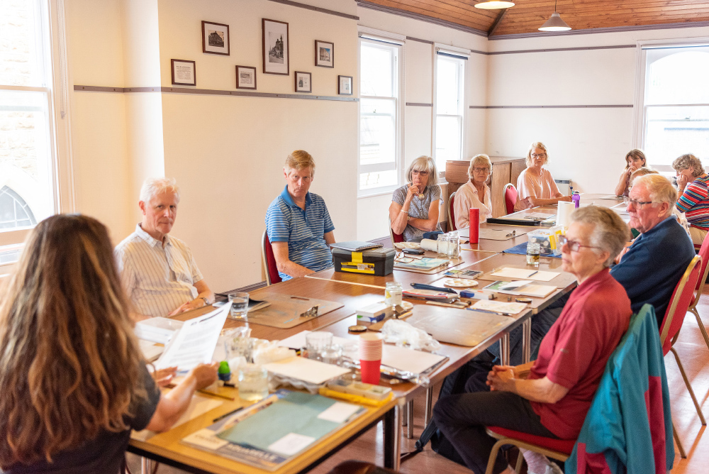 The Studio: 10 people sat around a long table in discussion. The walls are white, with framed prints on the wall and large sash windows. 