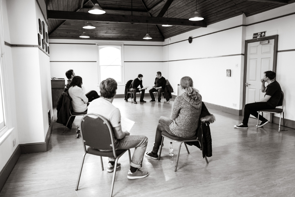 The Studio: A black and white image of 7 people taking part in an acting class. They are sat on chairs spaced around the room which features a vaulted ceiling.