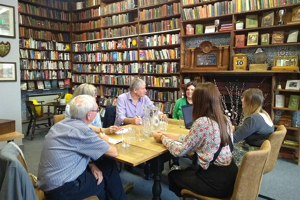 The Library: Six people sat at a table having a meeting, surrounded by floor-to-ceiling bookshelves.