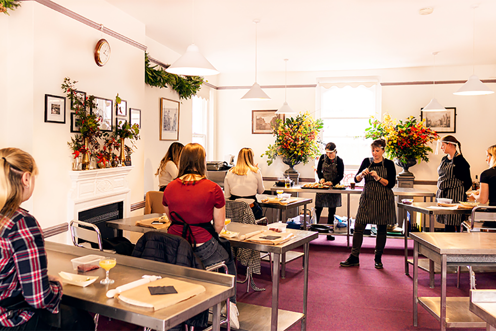 The Old Billiard Room being used for a cookery course. Groups of people are sat at wooden tables watching a demonstration at the front. The walls are covered with festive decorations and foliage.