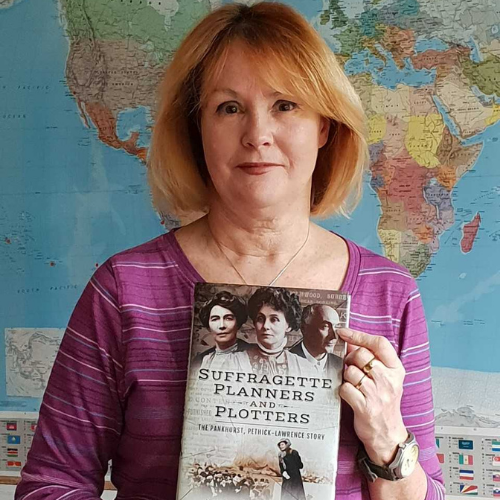 Photo of Kathy Atherton. She is wearing a purple top and holding a copy of her book, 'Suffragette Planners and Plotters', stood in front of a world map.