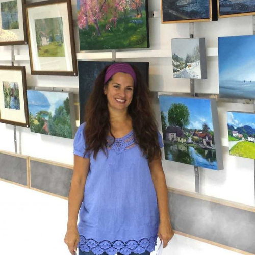 Picture of Songul Yilmaz Meier. She is wearing a blue top and purple hairband, stood in front of display of her paintings.