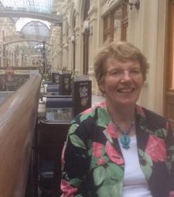 Institute tutor, Susan Purcell, sat smiling wearing a black, pink and green floral jacket, blue necklace and glasses.