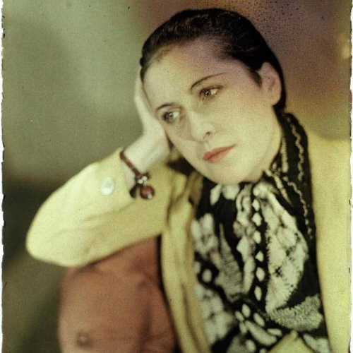 International Women's Day: Photo of Dora Maar. She is captured sitting down, looking away from the camera, with her hand resting on her head. She is wearing a yellow jacket and black and white floral top. 