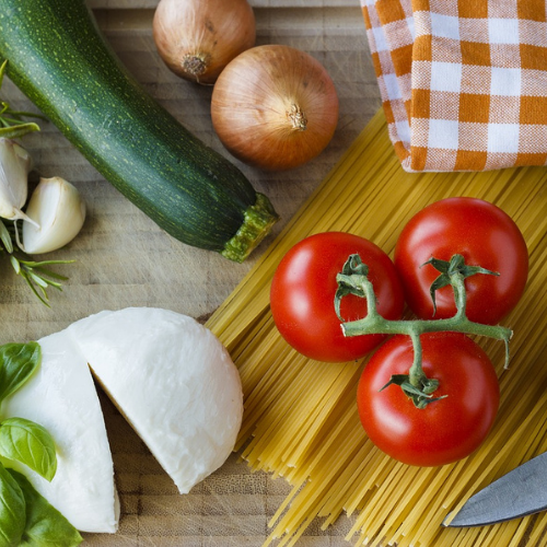 Flat-lay of cooking ingredients including tomatoes, spaghetti, mozzarella, courgette and onions. 