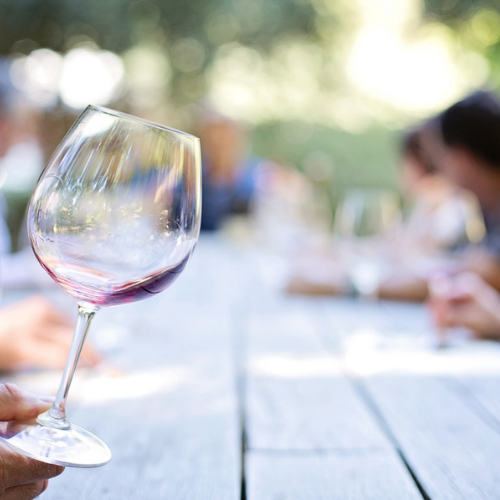 5 events to look out for: Summer Wine Tasting. Image of a hand holding a wine glass in the foreground. In the background there is a wooden table with guests seated around it. 