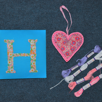 A blue square embroidered with a floral letter H; a pink heart embroidered with red and blue flowers; purple, white and pink thread.