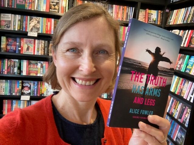 Alice Fowler standing in front of bookshelves, holding a copy of her short stories collection, The Truth Has Arms And Legs