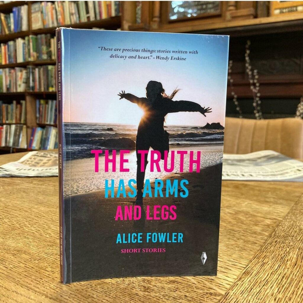 Close-up of Alice Fowler's short stories collection, The Truth Has Arms and Legs. The cover is a silhouette of a figure running on the beach. The book is  placed on a table with bookshelves in the background.