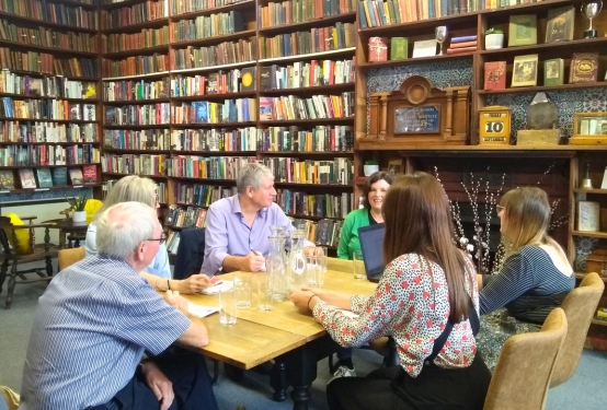 Six peopl sat around a table having a meeting in a library with floor-to-ceiling bookshelves