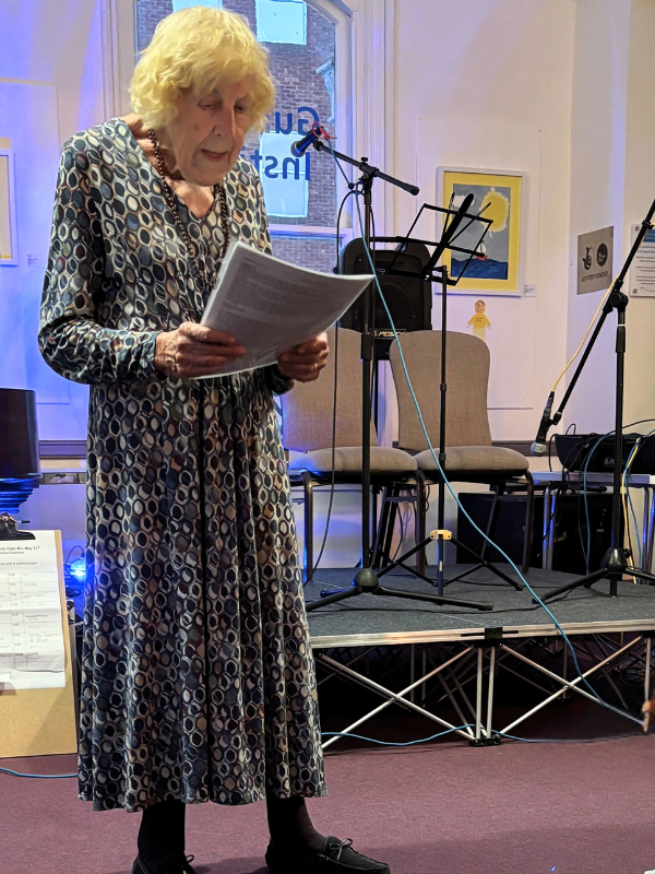 Open Mic Nights: a lady with short blonde hair and a patterned dress stood  reading poetry