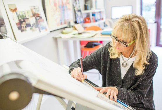 Woman with long blonde hair and a grey cardigan, sat in a studio sketching at an easel.